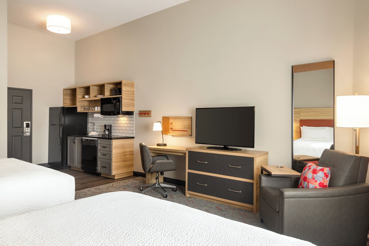 A hotel room with a bed, a desk with a computer, a television, a chair, a small kitchenette, and a mirror.