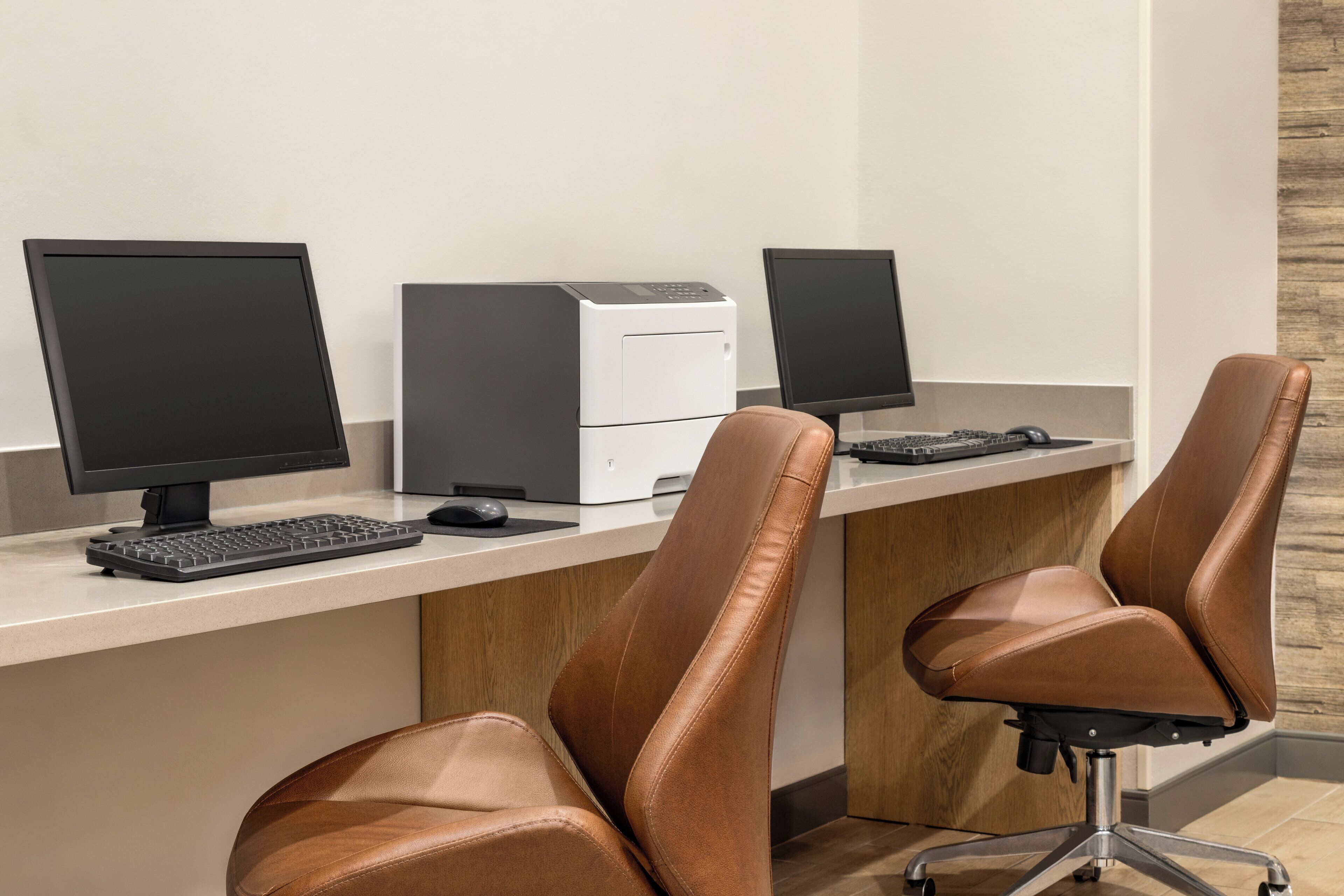 A modern office space with three desks, each equipped with a computer monitor, keyboard, and mouse. The desks are arranged in a row with brown leather chairs in front of them.