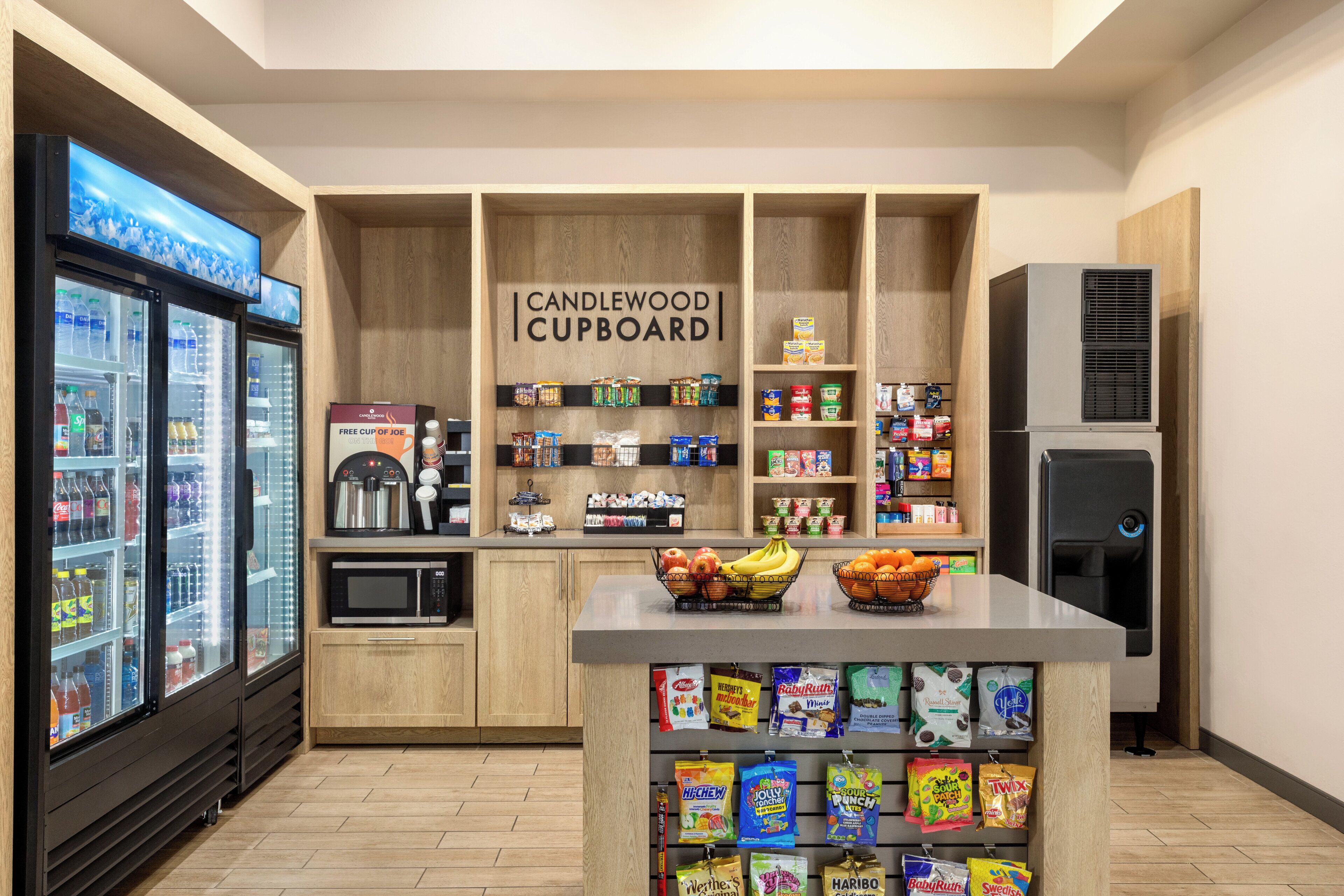 A kitchenette area with a microwave, refrigerator, and a coffee machine. There are shelves with snacks and a sign that reads "CANDLEWOOD CUPBOARD".