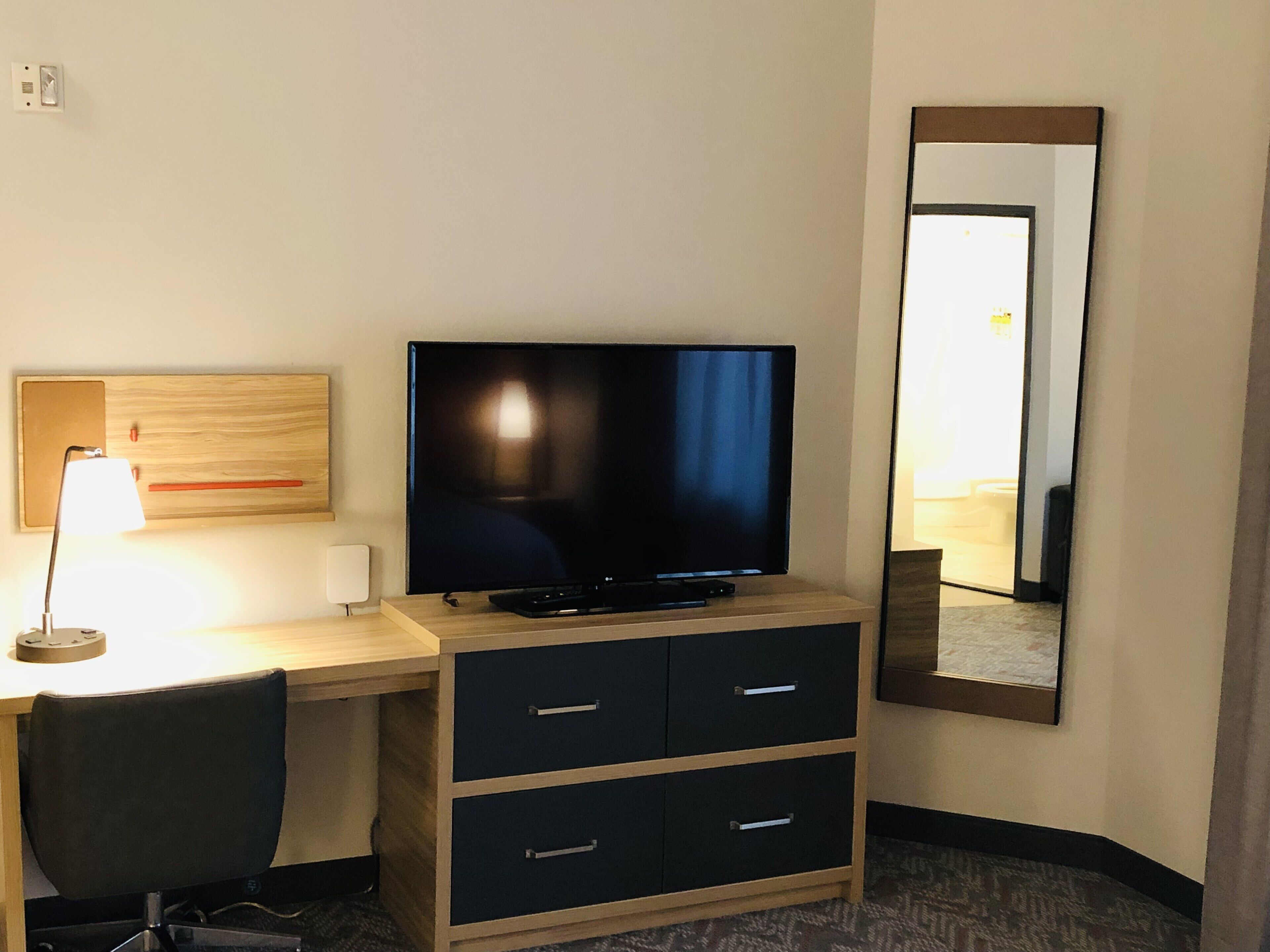 A hotel room with a desk, a television, a chair, a mirror, and a lamp.