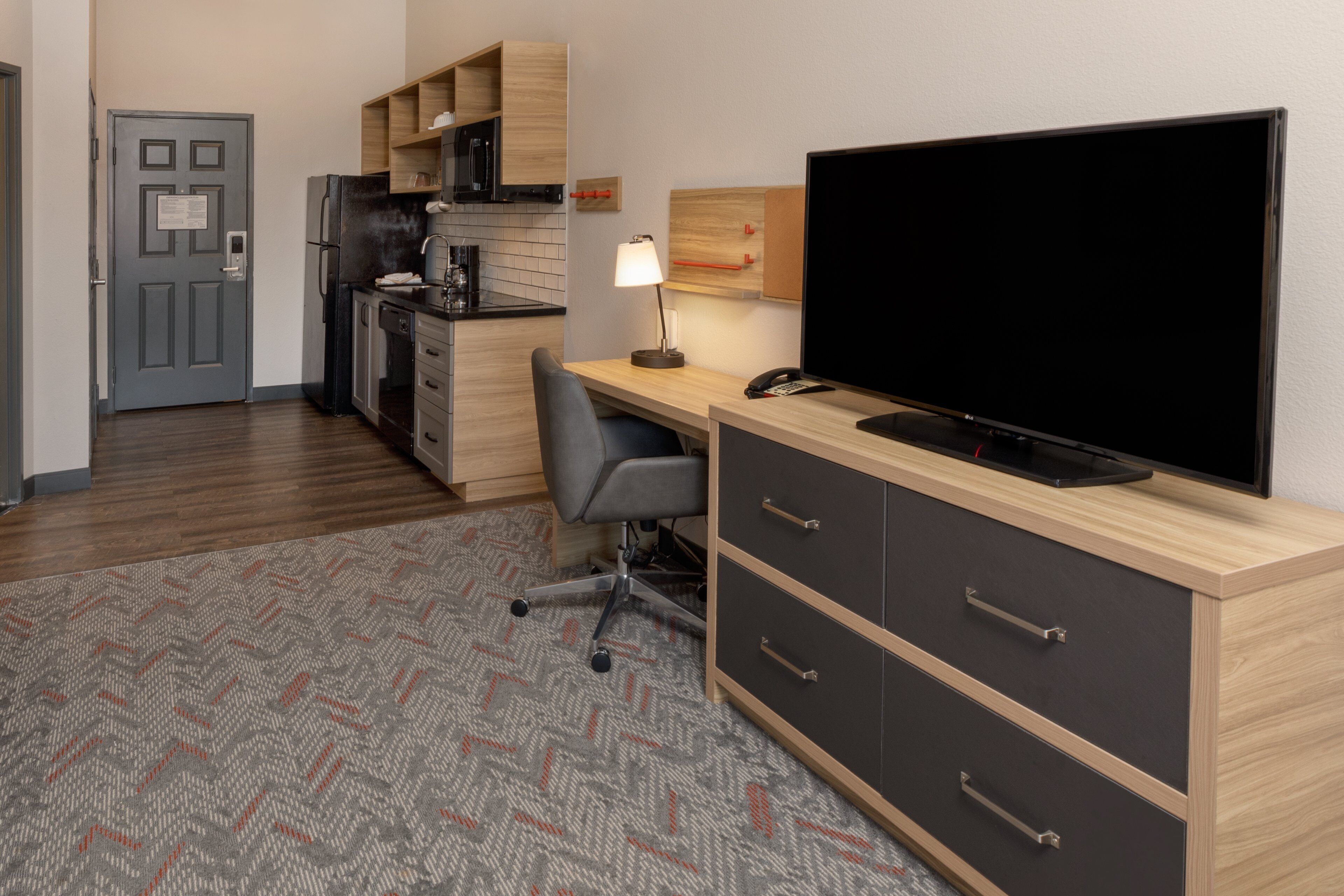 Habitación de hotel compacta con kitchenette, escritorio, televisor de pantalla plana, una silla y alfombra estampada.