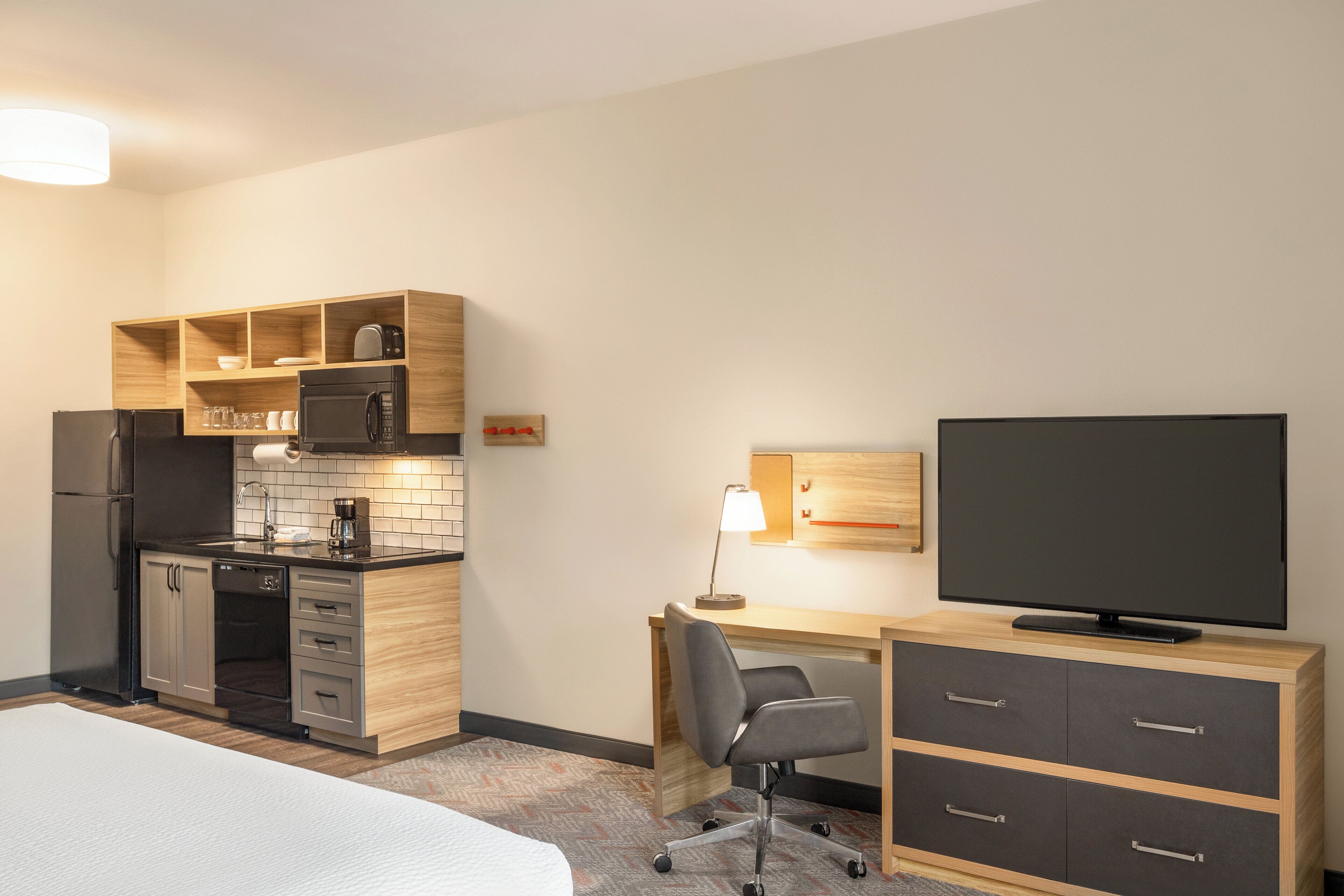 A compact hotel room with a kitchenette, a bed, a desk with a chair, and a television.