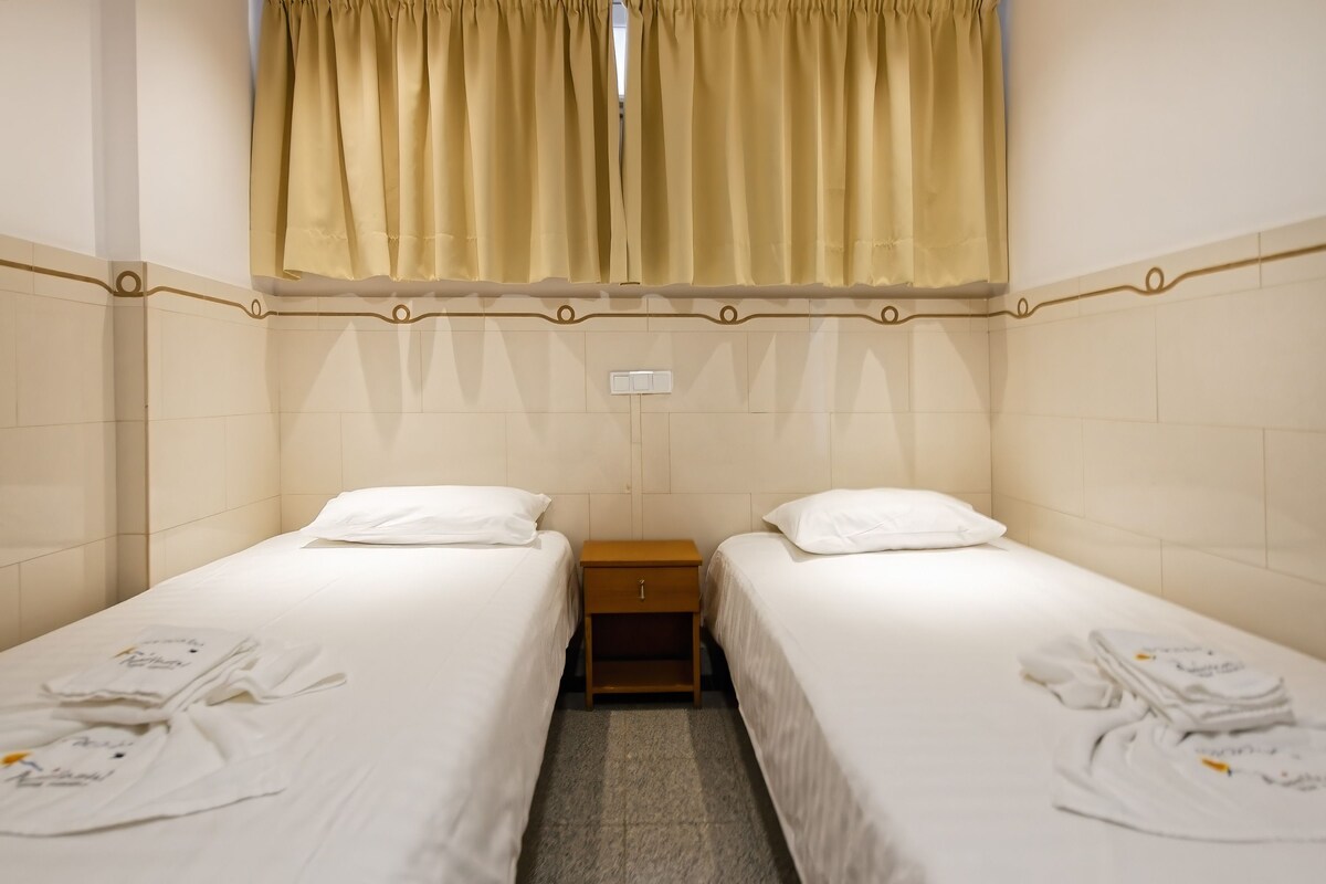 Habitación de hotel con dos camas individuales, una mesita de noche de madera y una ventana con cortinas color beige.