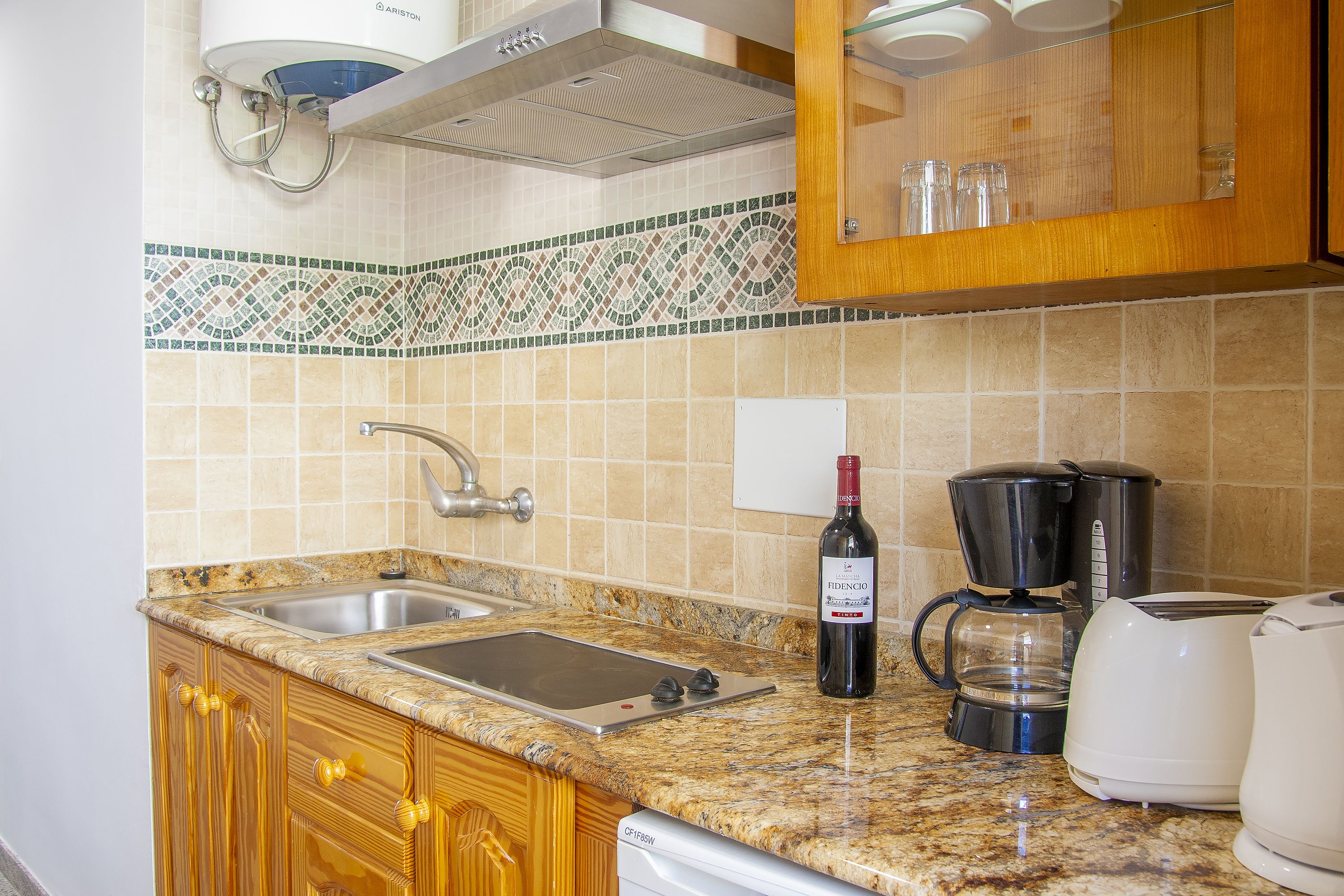 A kitchen with a granite countertop, stainless steel sink, wooden cabinets, a coffee maker, and a wine bottle.