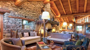 Un interior rústico con paredes de piedra, vigas de madera, una chimenea de piedra y varios asientos, incluyendo un sofá, sillas y un banco.