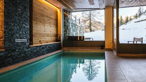 Una piscina interior moderna con paredes de piedra y paneles de madera, grandes ventanales que ofrecen vistas a un área exterior nevada.