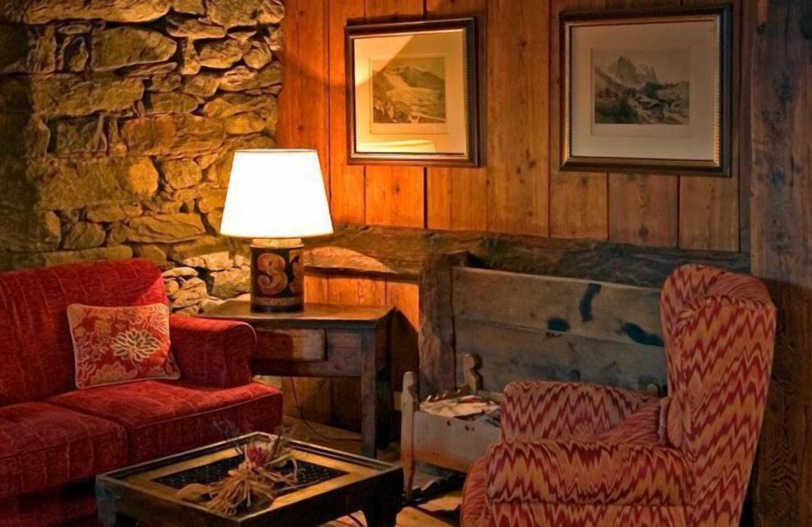 Une pièce chaleureuse avec un mur en pierre, un canapé rouge, un fauteuil à motifs et une table avec une lampe.