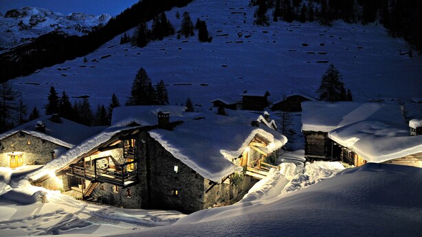 Un refugio de montaña cubierto de nieve con fachada de madera y numerosas ventanas, rodeado de árboles nevados y un cielo nocturno despejado.