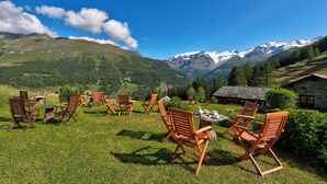 Un área de estar al aire libre con vistas panorámicas a un paisaje montañoso con picos nevados y abundante vegetación, incluyendo sillas y mesas de madera.