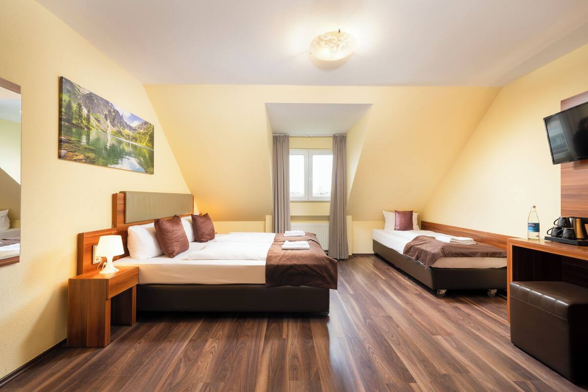 Une chambre d’hôtel avec un grand lit, un lit plus petit, un bureau avec une télévision, un tableau au mur et une fenêtre avec des rideaux.