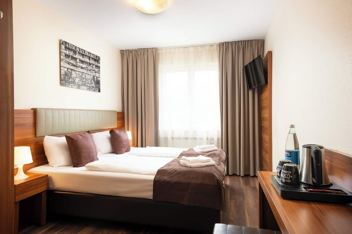 Une chambre d’hôtel avec un lit, une table de chevet avec une lampe, un téléviseur à écran plat fixé au mur et une fenêtre avec des rideaux.