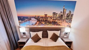 Ein Hotelzimmer mit einem großen Bett, zwei Kissen, einem Kopfteil, einem Nachttisch mit Lampe und Blick auf eine Stadtlandschaft bei Sonnenuntergang.