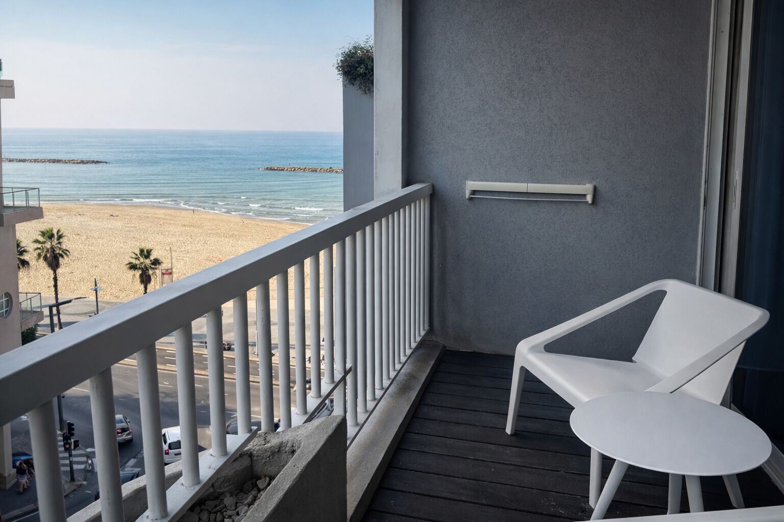 En balkon med hvid stol og bord, der kigger ud over en strand med palmer og en mol.