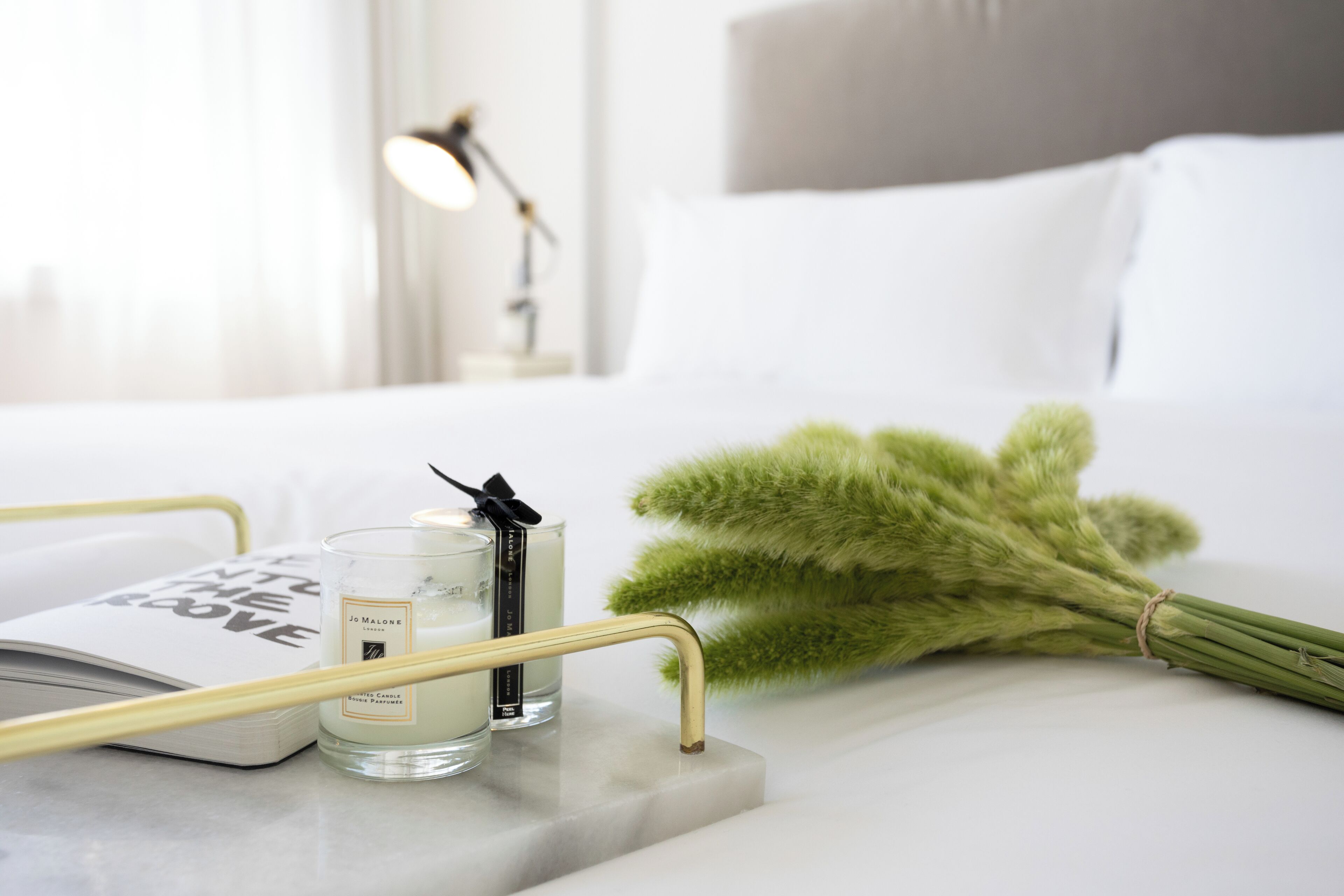 A neatly arranged bedroom with a bed, a bedside table with a candle and a book, and a bouquet of green pampas grass.