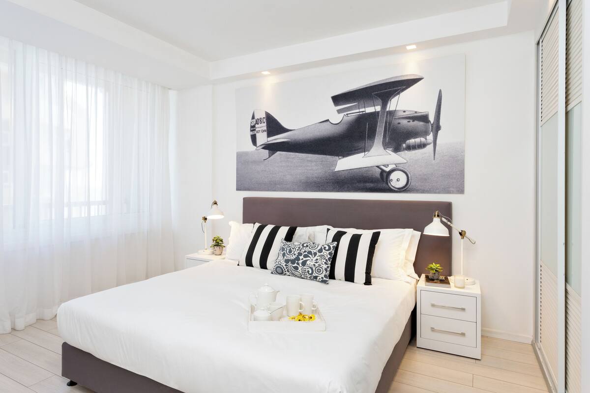A modern hotel room with a large bed, a nightstand with a lamp, a framed airplane picture, and a tray with a teapot and cups.