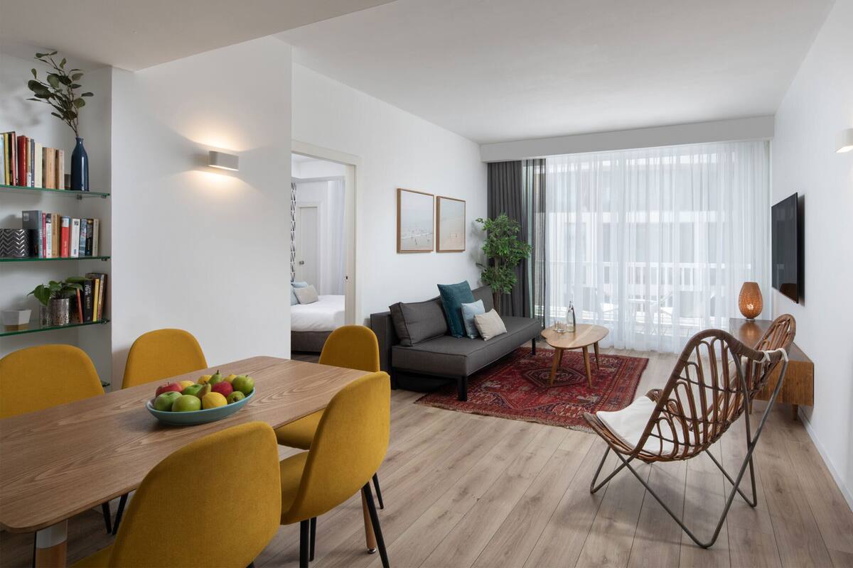 A modern living room with a sofa, dining table, and chairs. There is a bookshelf, a plant, and a TV mounted on the wall.