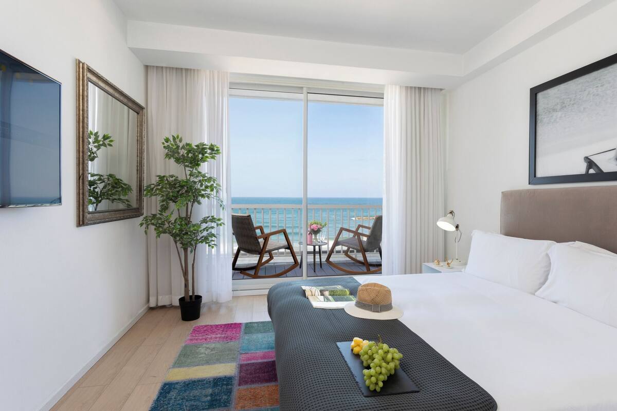 A hotel room with a large bed, a view of the sea, a balcony with chairs, and a plant.