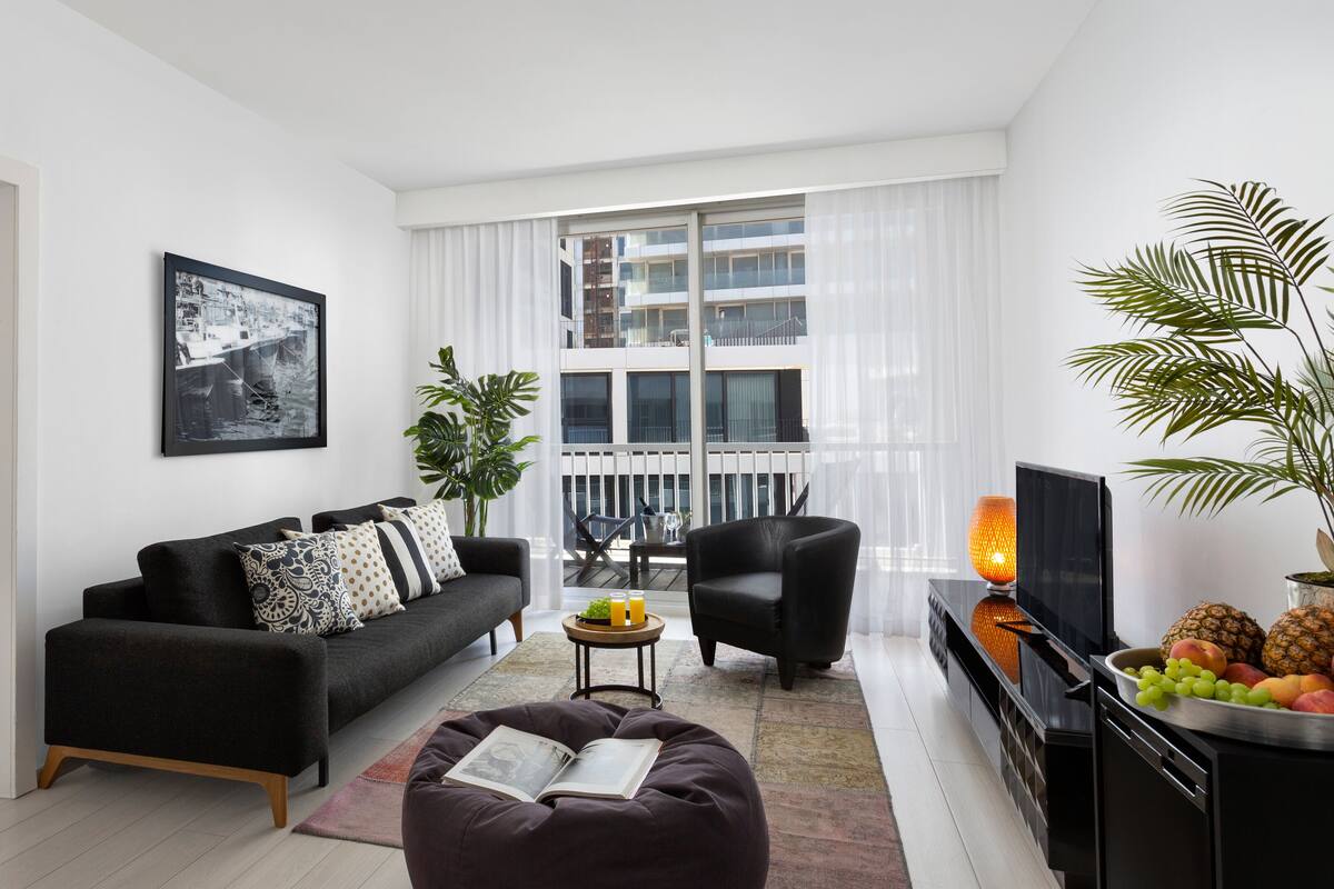 A modern living room with a sofa, a coffee table, a TV, and a balcony with a view of buildings.