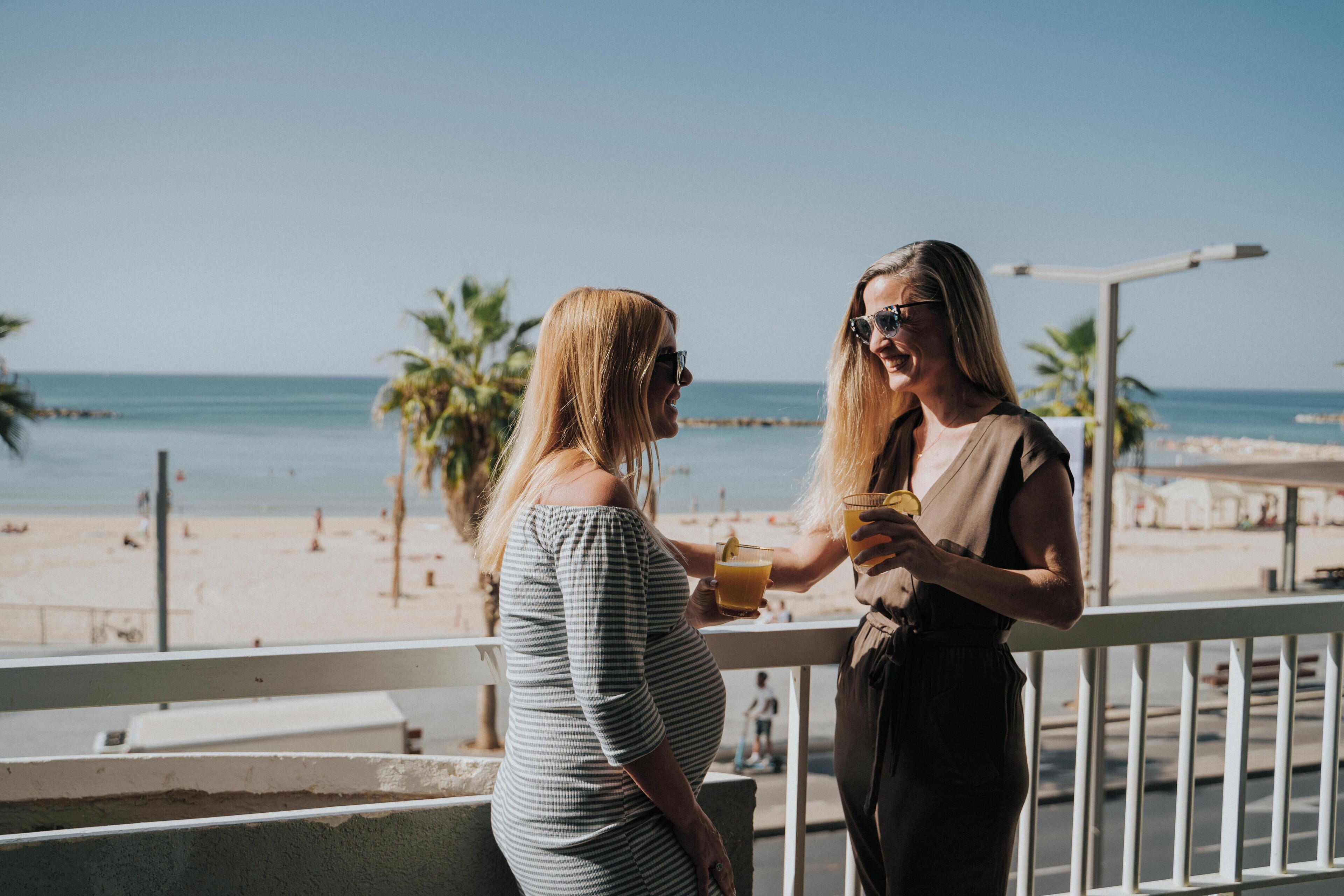 Two women on a balcony overlooking a beach with palm trees and the ocean.