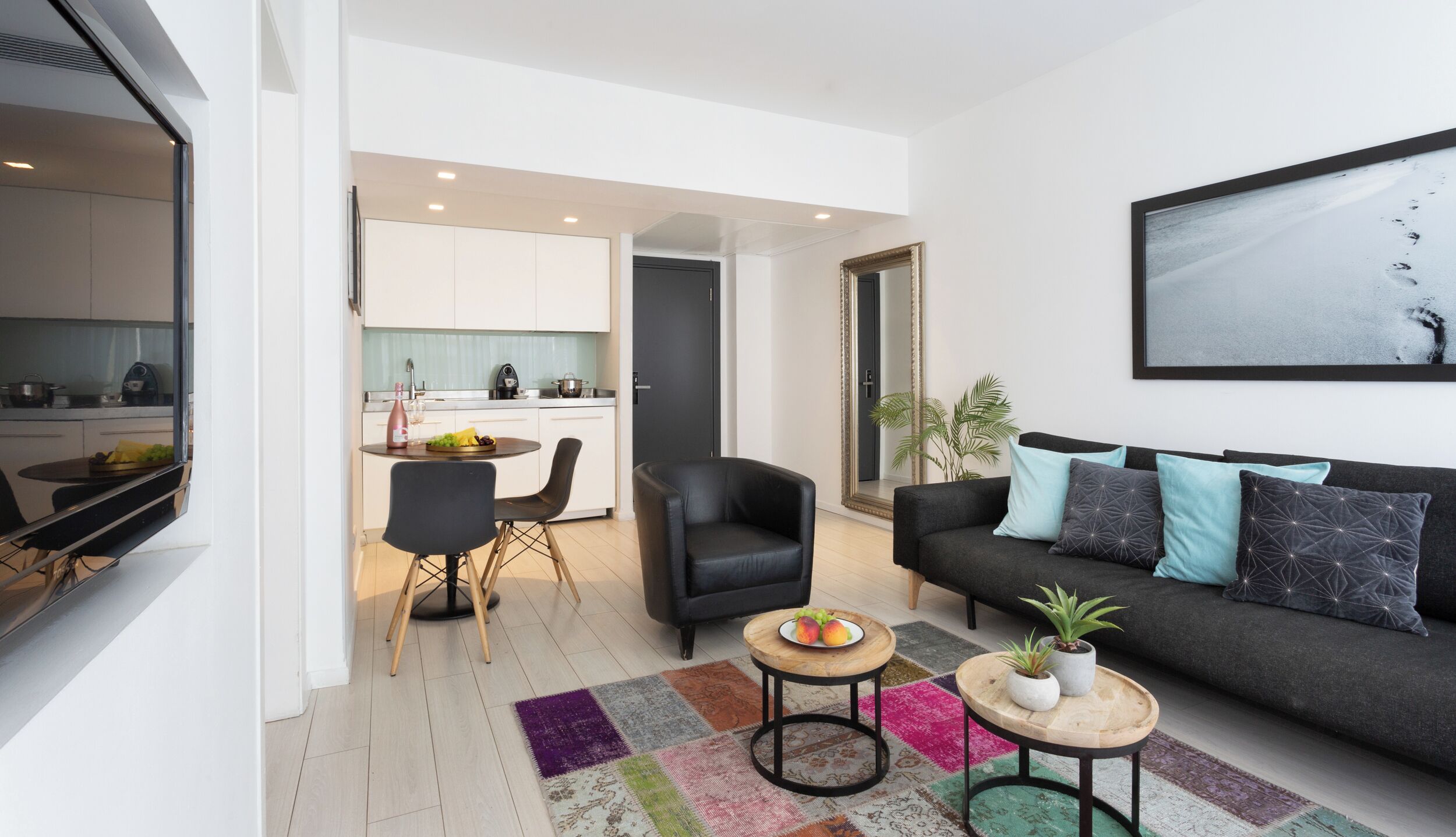 A modern living room with a black sofa, a dining area with a round table, and a kitchen visible through an open door.