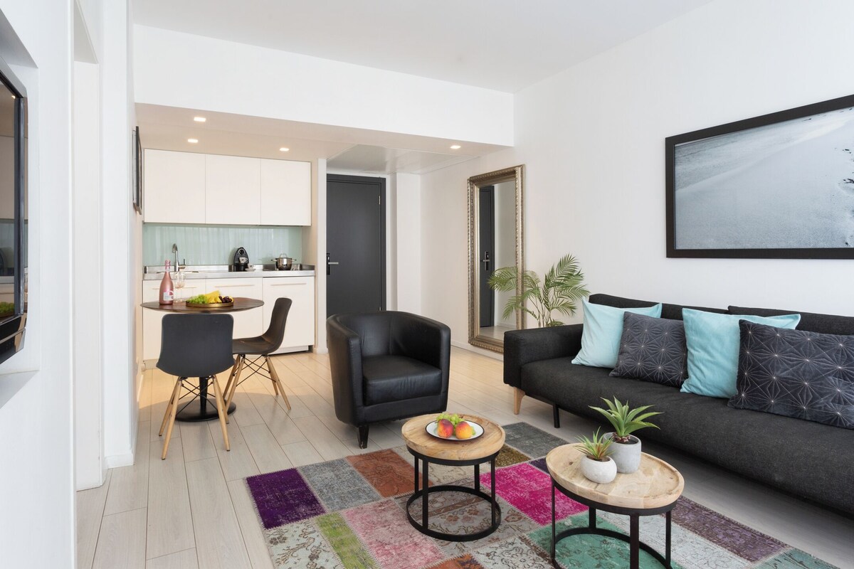 A modern living room with a black sofa, a dining area with a round table, and a kitchen visible through an open door.