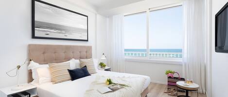 Un dormitorio moderno con una cama grande, una mesita de noche con una lámpara, una ventana con vista al mar y una pequeña mesa redonda con una planta.
