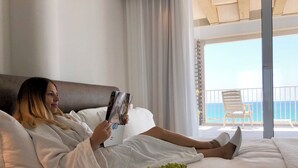 A person lying on a bed in a hotel room, reading a magazine, with a bowl of grapes and a view of the sea through the balcony. - Sea Executive Suites (Tel Aviv)