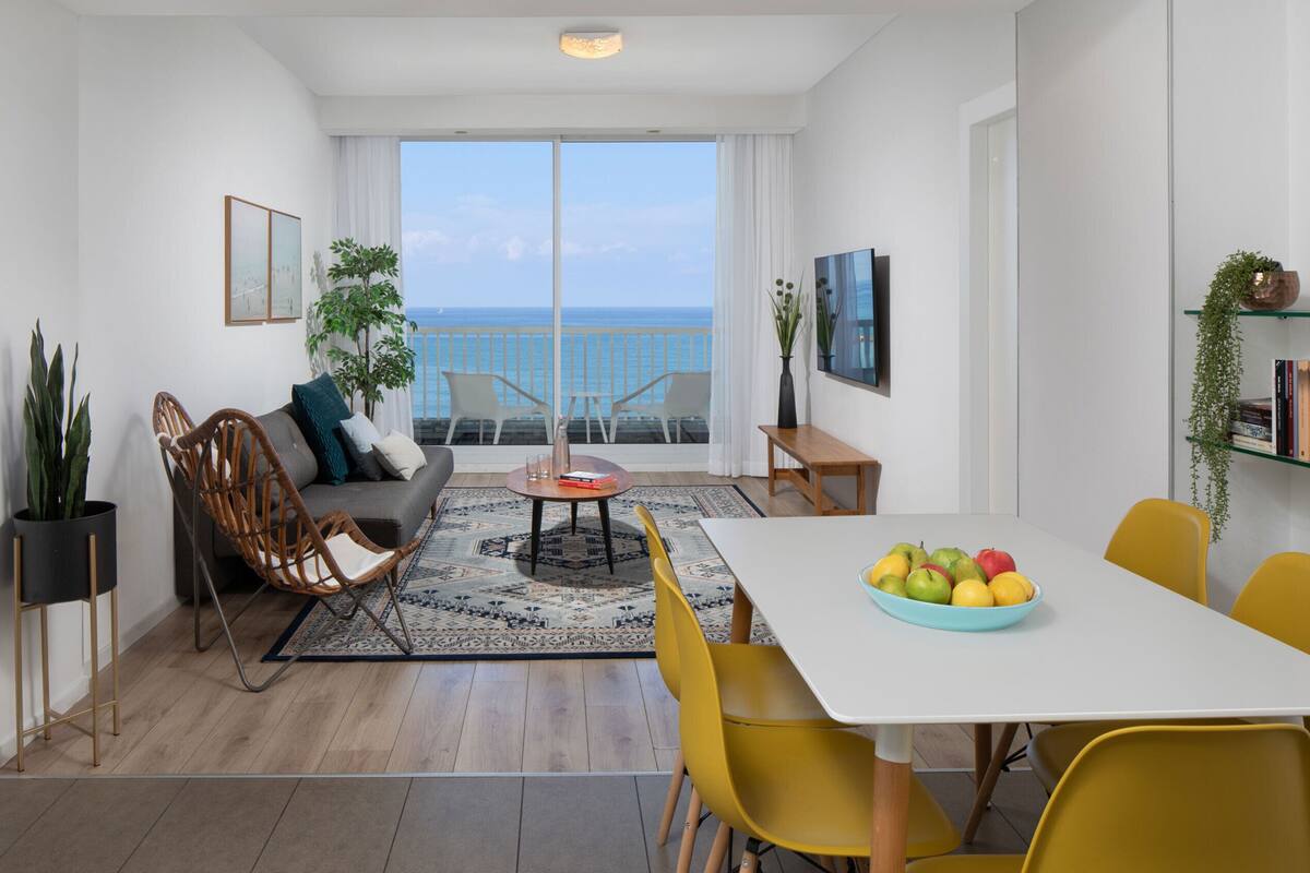 A modern living room with a sofa, chairs, a dining table with fruit, and a balcony with a view of the sea.