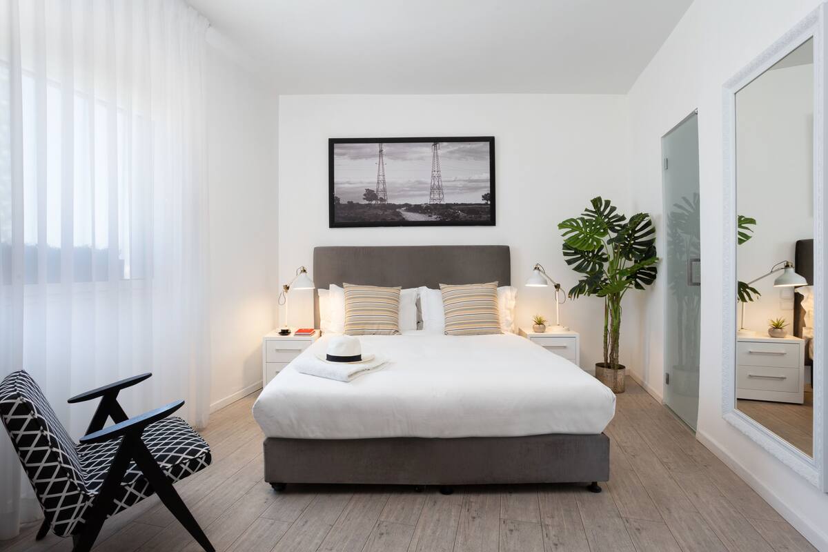 A modern bedroom with a large bed, bedside tables, a nightstand, a chair, a potted plant, and a large mirror.