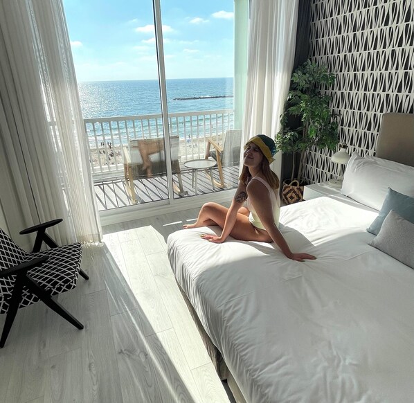 A bedroom with a large window overlooking a beach, a bed, a chair, and a small table.