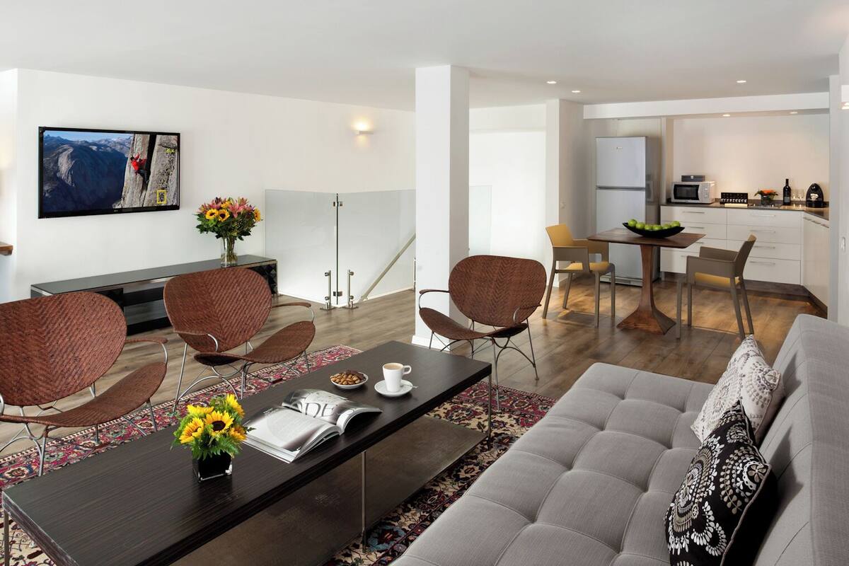 A modern living room with a sofa, chairs, a coffee table, and a TV. There is a kitchen area with a dining table and chairs.