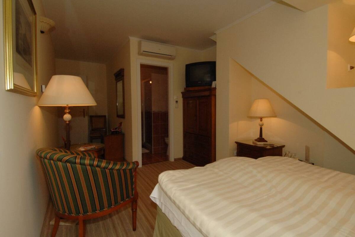 Una habitació d'hotel amb un llit, una cadira, una taula de treball amb una làmpada, una televisió damunt un moble i un aire condicionat.