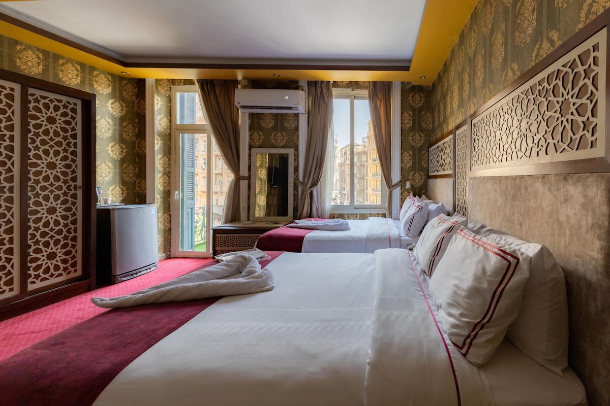 A hotel room with two beds, a red carpet, a decorative wall, and a view of buildings through the window.