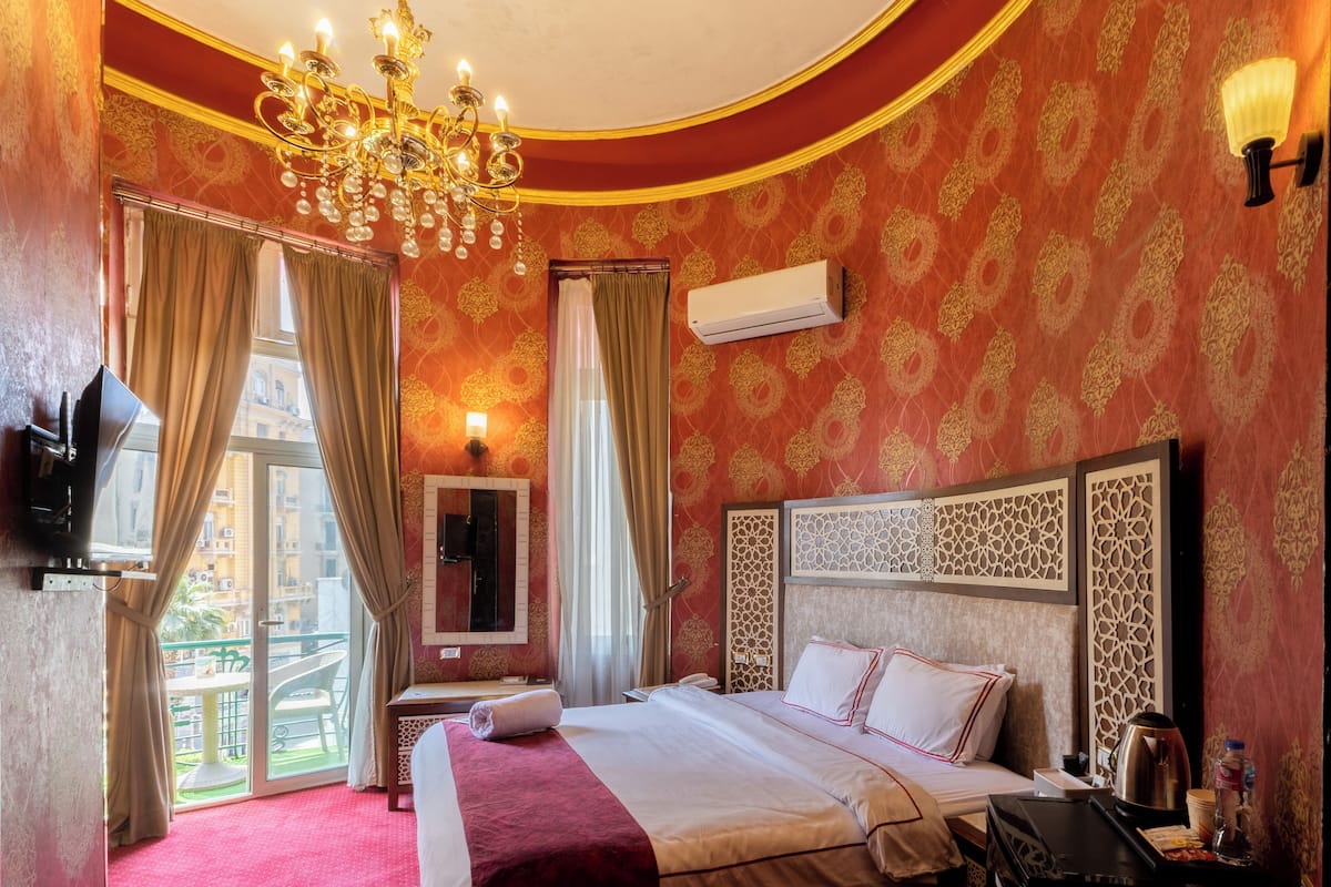 A hotel room with a large bed, a chandelier, a flat-screen TV, and a balcony with curtains.