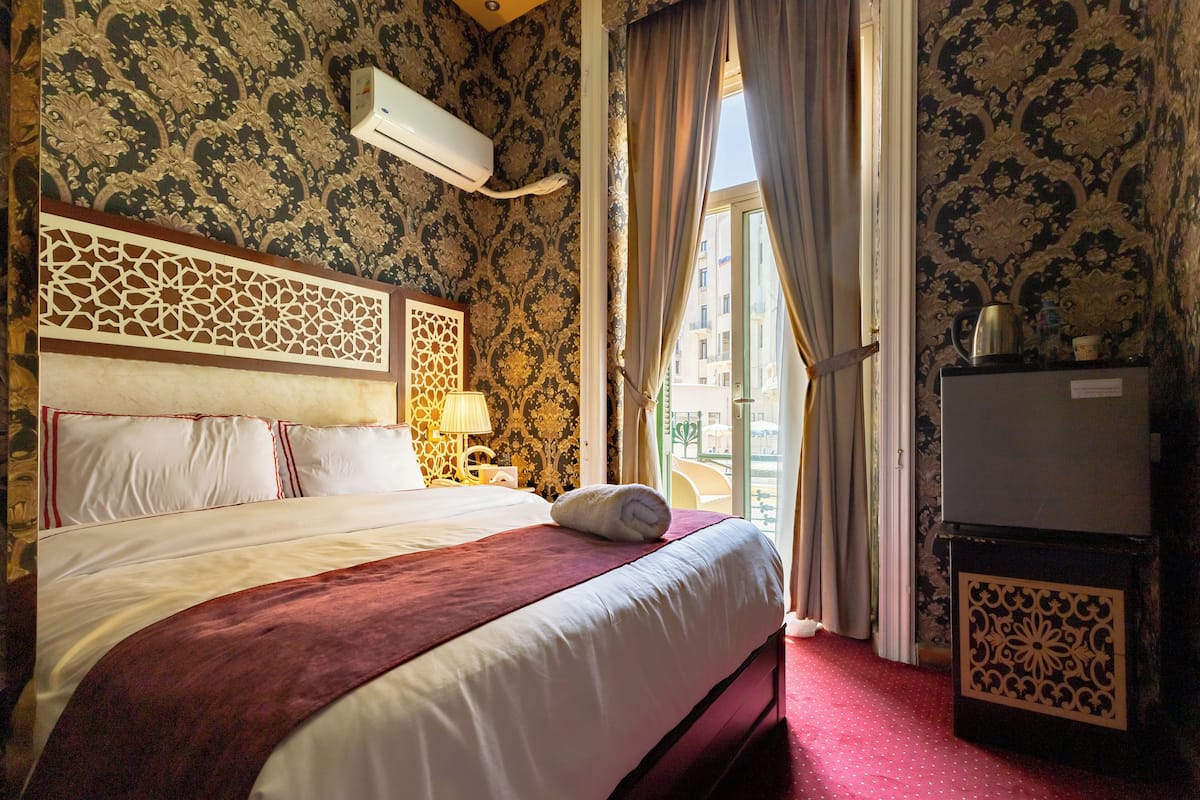 A hotel room with a large bed, a decorative headboard, a bedside table with a lamp, and a view of the city through the window.