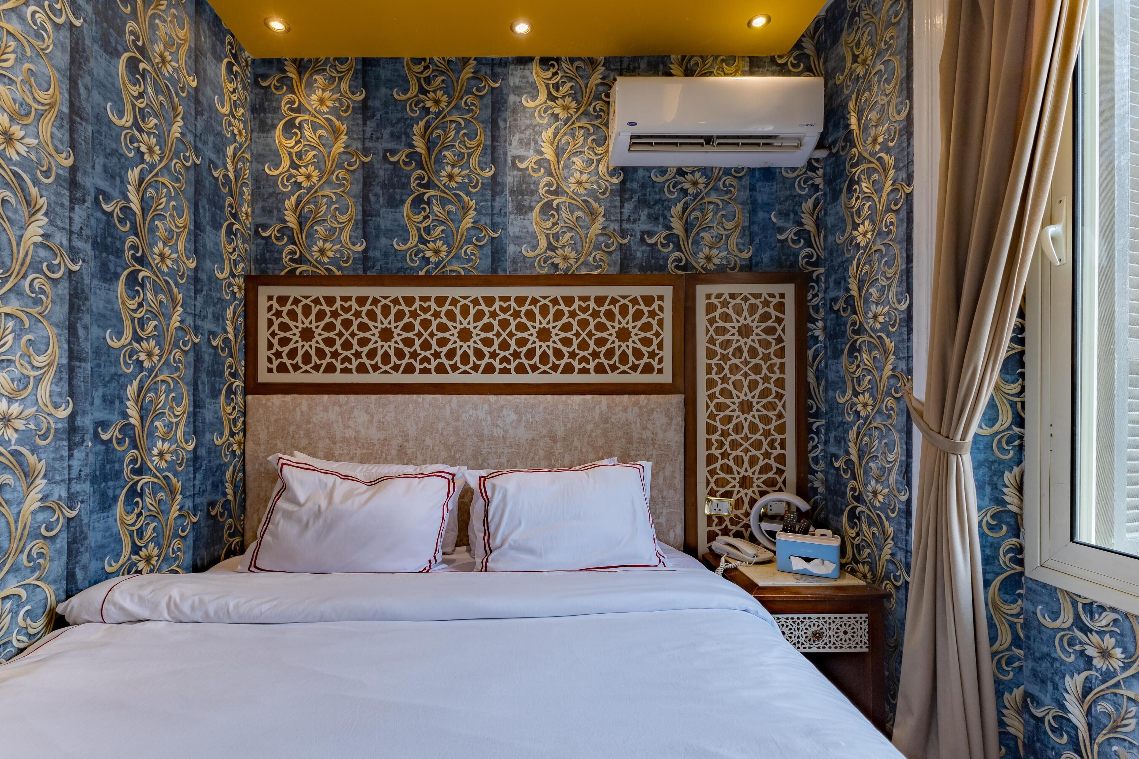 Una habitación de hotel con cama, mesita de noche, equipo de aire acondicionado y ventana con cortinas.