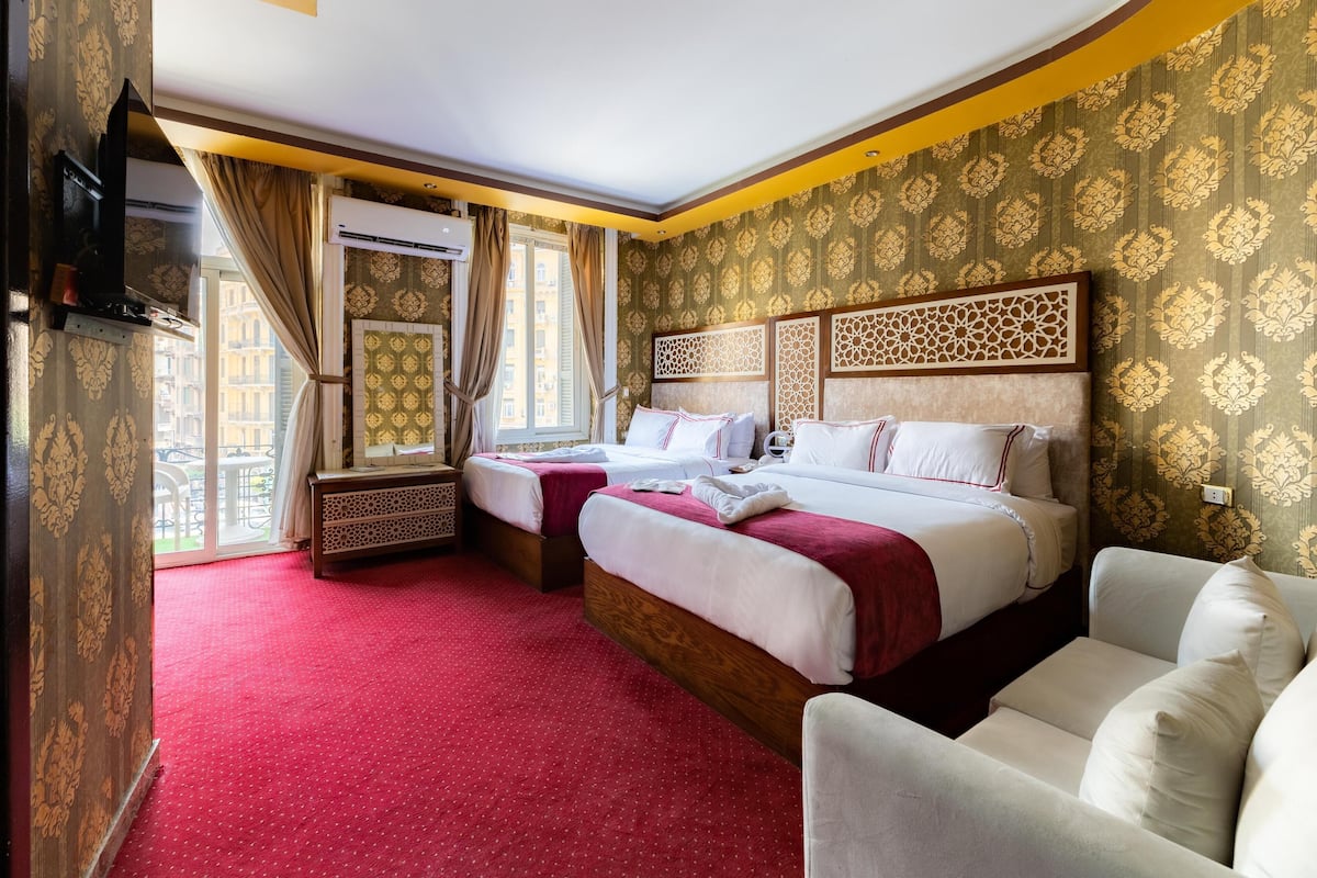 A hotel room with two beds, a red carpet, patterned wallpaper, and a view of buildings outside.