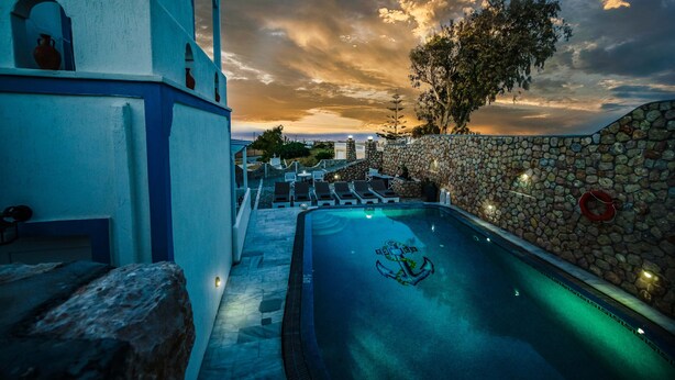 A swimming pool with an anchor logo, stone walls, and lounge chairs. - Stavros Villas (Santorini)