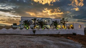 A modern building with multiple arched windows and a white facade, surrounded by palm trees and a gravel driveway. - Stavros Villas (Santorini)