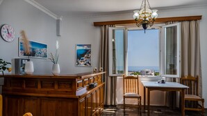 A room with a wooden cabinet, a dining table with chairs, a chandelier, and a large window with a view of the sea. - Stavros Villas (Santorini)