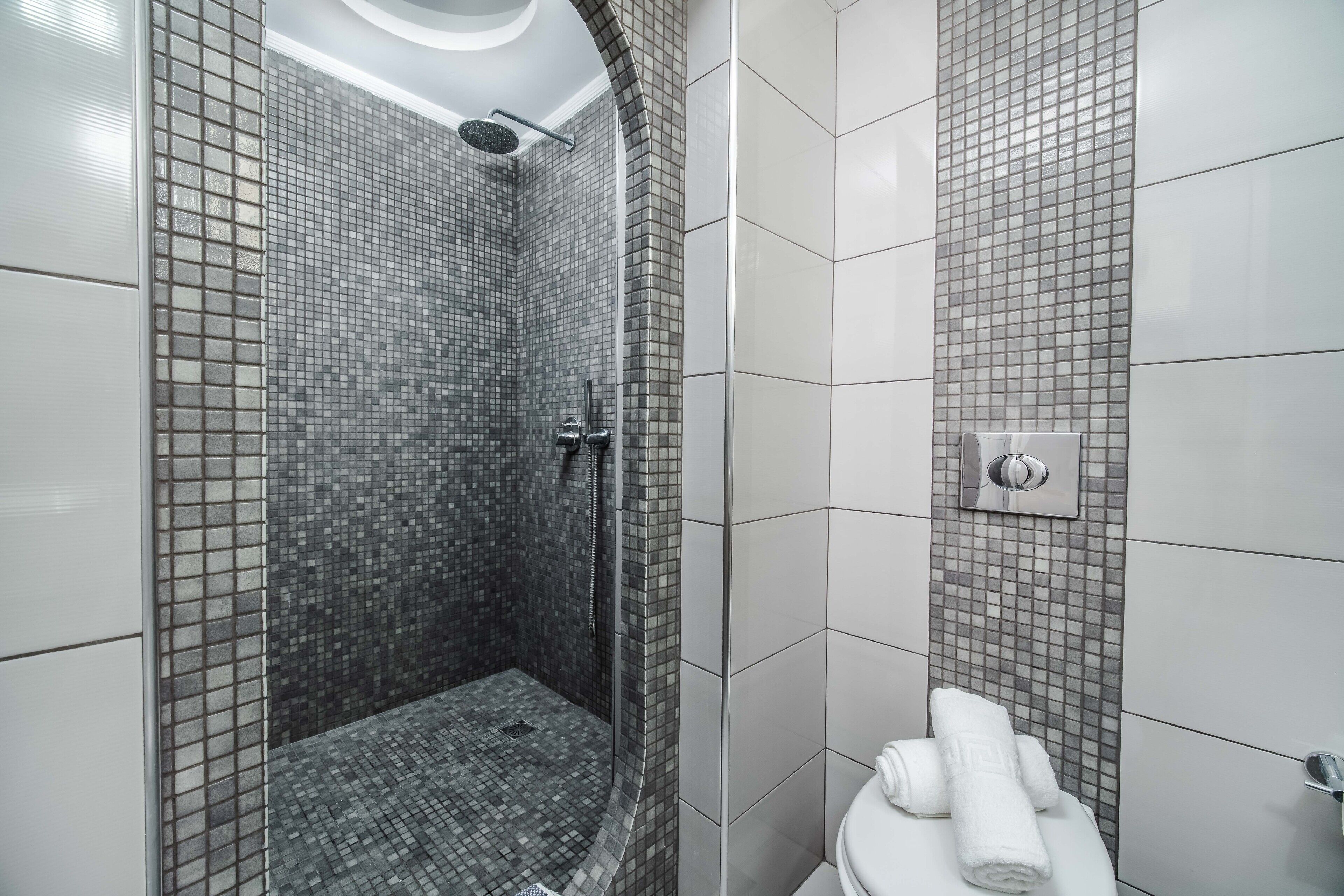 A modern bathroom with a shower area tiled in a mosaic pattern, a toilet with a towel rack, and a wall-mounted light fixture.
