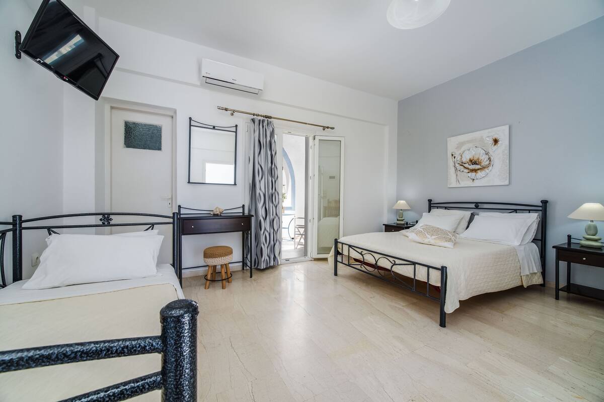 A bedroom with two beds, a desk, a chair, a small stool, a bedside table with a lamp, and a wall-mounted TV.