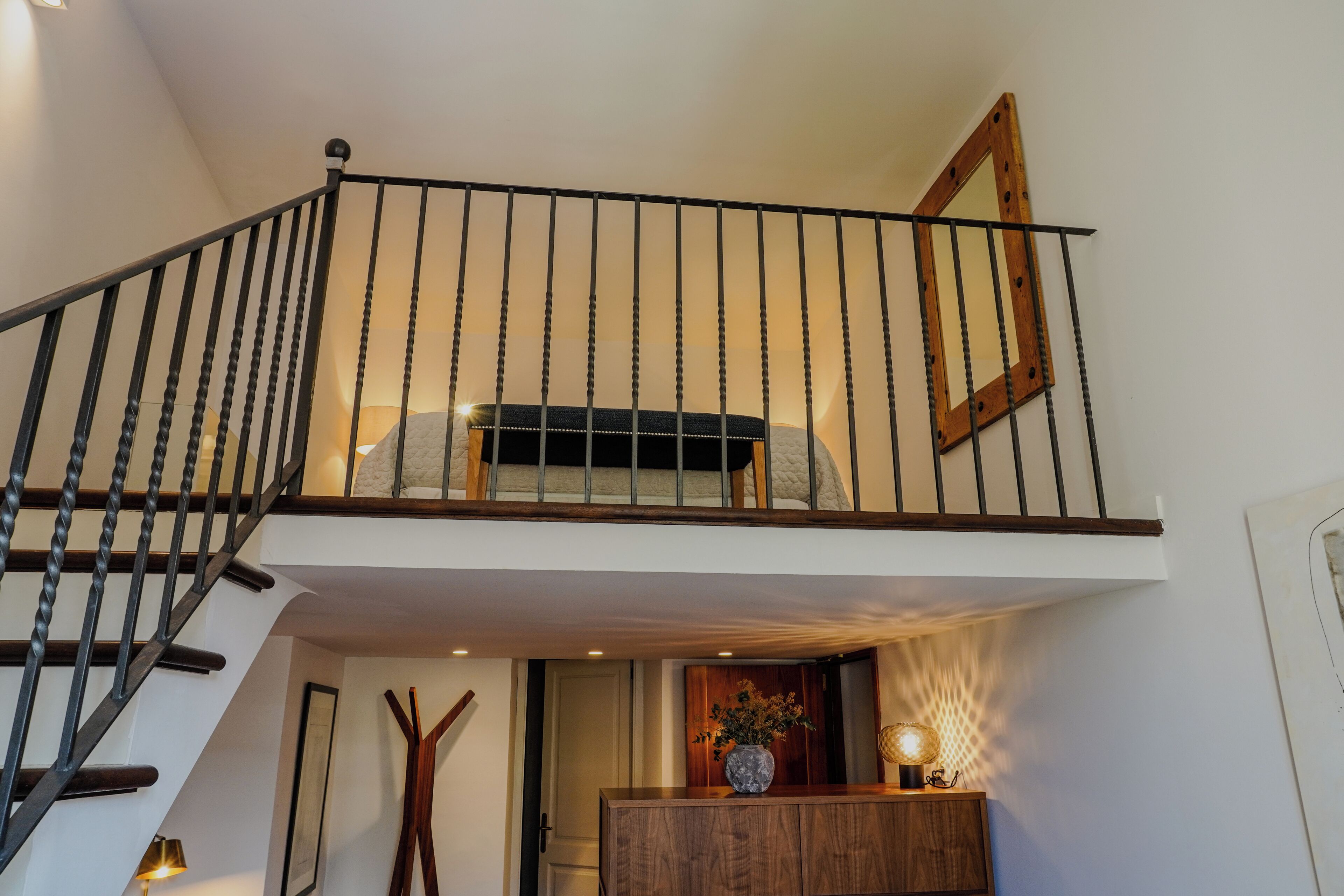Una escalera conduce a un dormitorio en lo alto, con barandilla de madera, una mesita de noche con lámpara y un espejo enmarcado en la pared.