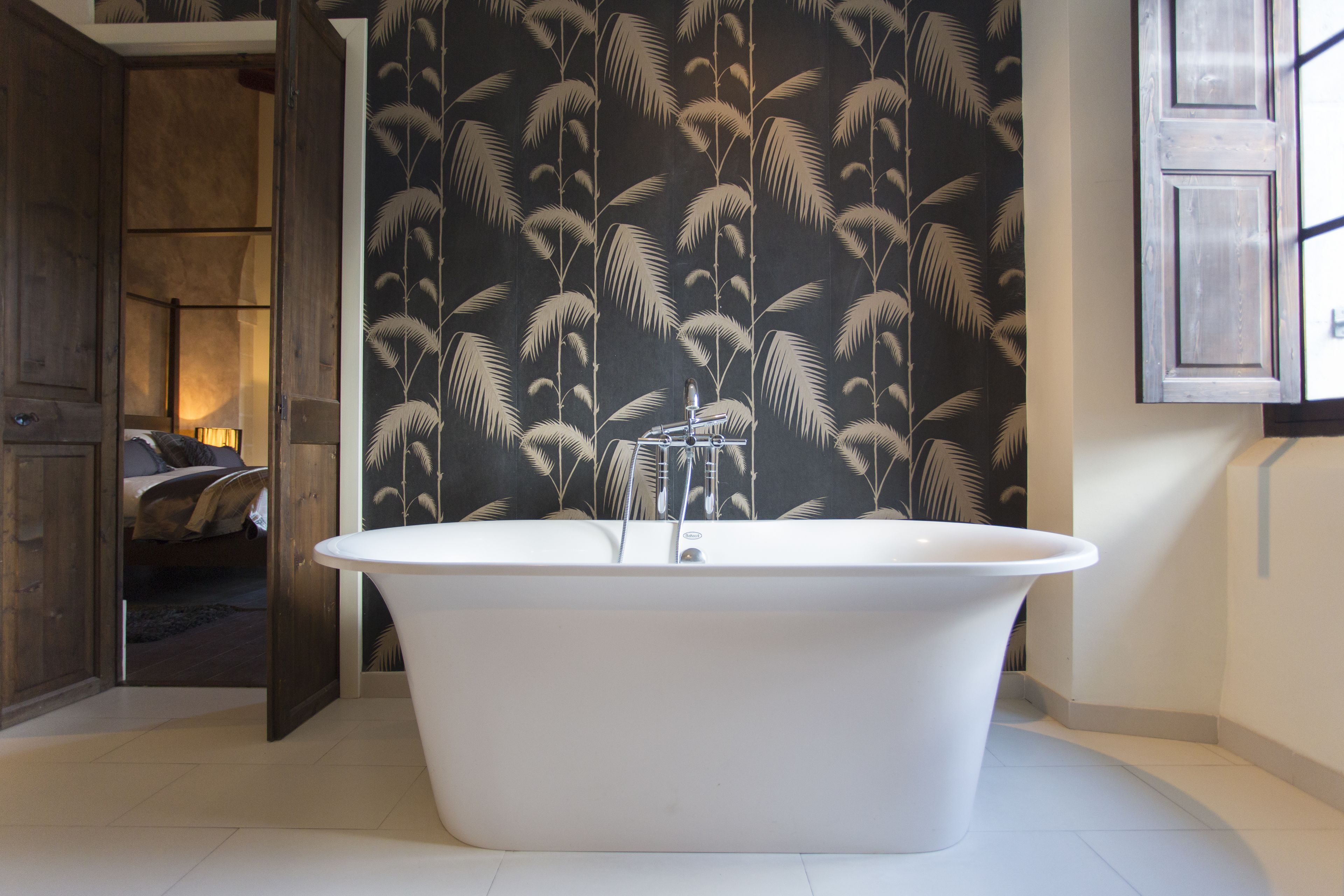 A modern bathroom with a freestanding bathtub, a wooden door leading to another room, and a patterned wallpaper.