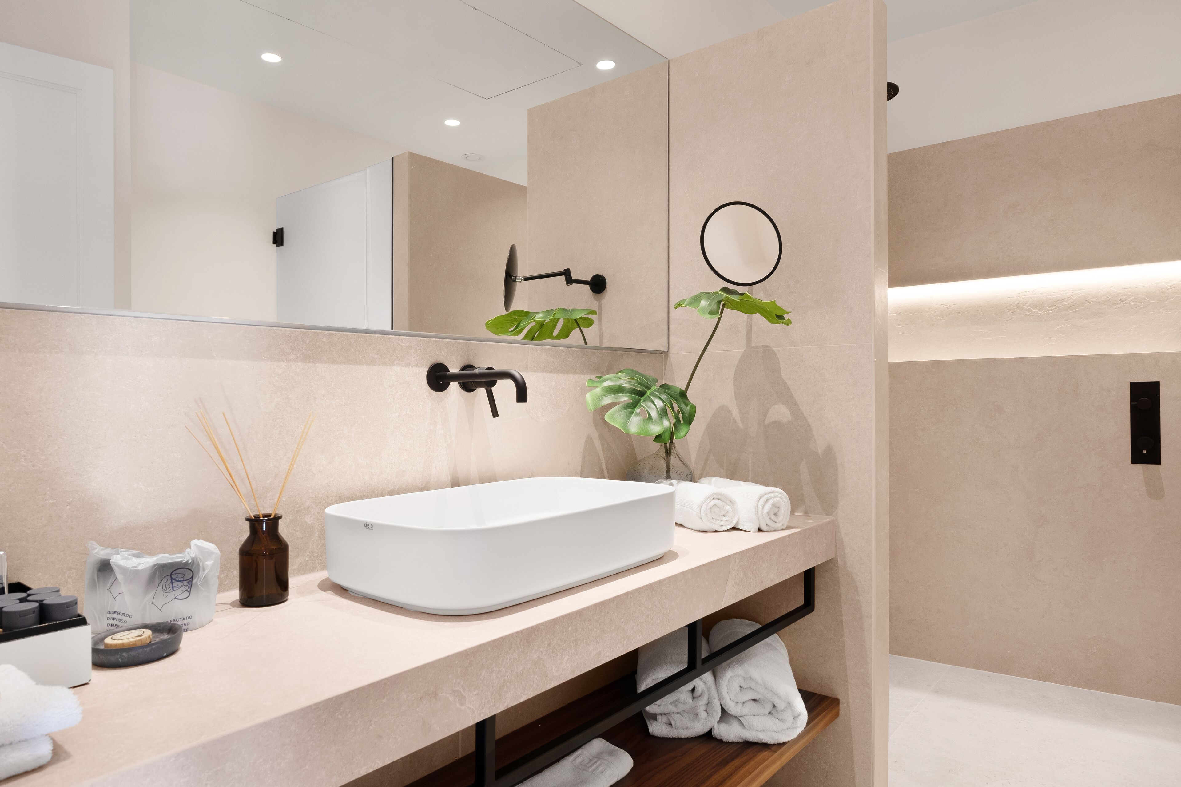 Un baño moderno con un espejo grande, un lavabo blanco, una planta y toallas dobladas con cuidado.