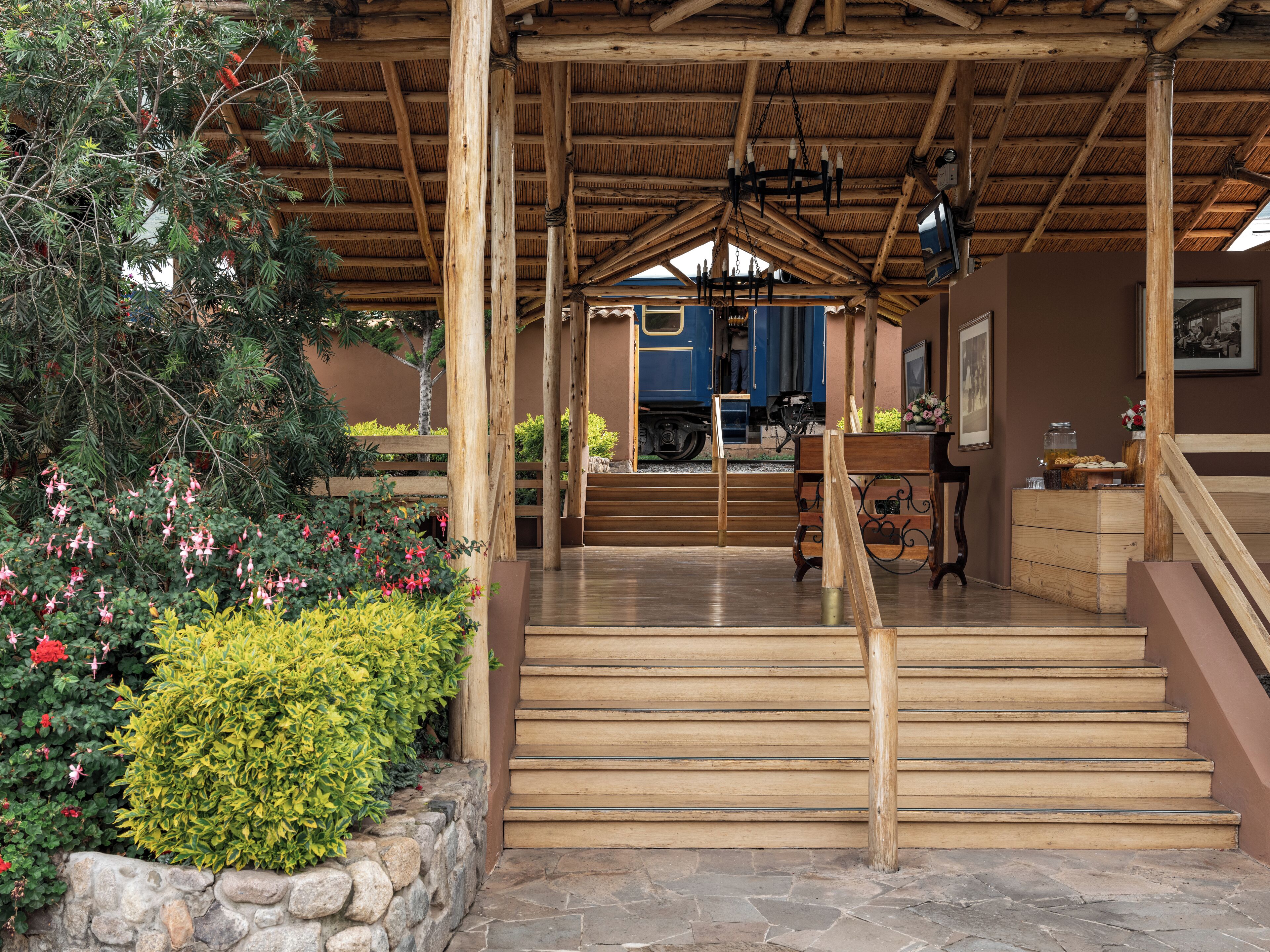 A wooden covered outdoor area with steps leading to a blue door, surrounded by greenery and stone walls.