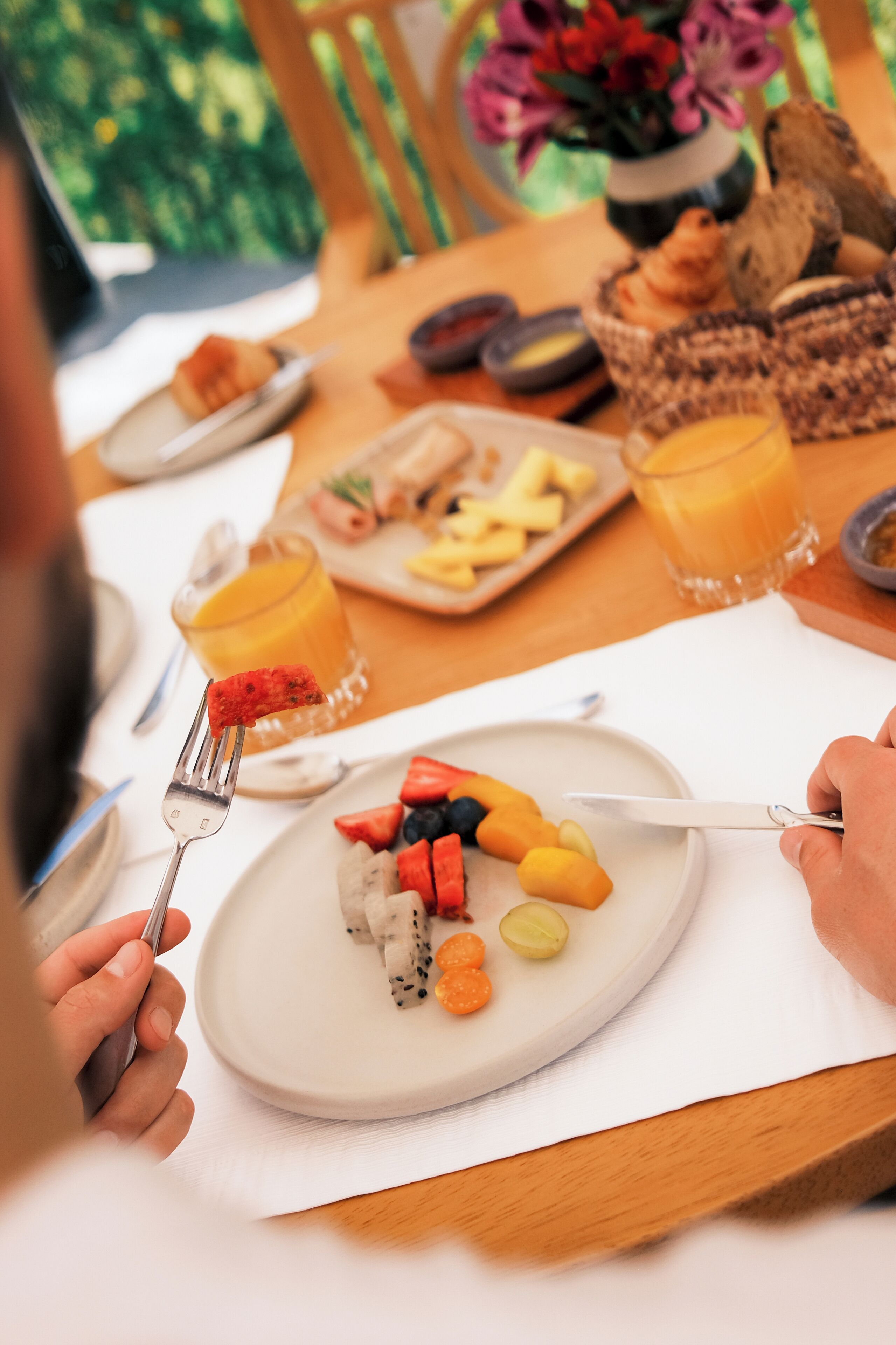 A dining table with a plate of fruit, a glass of orange juice, and a basket of bread.