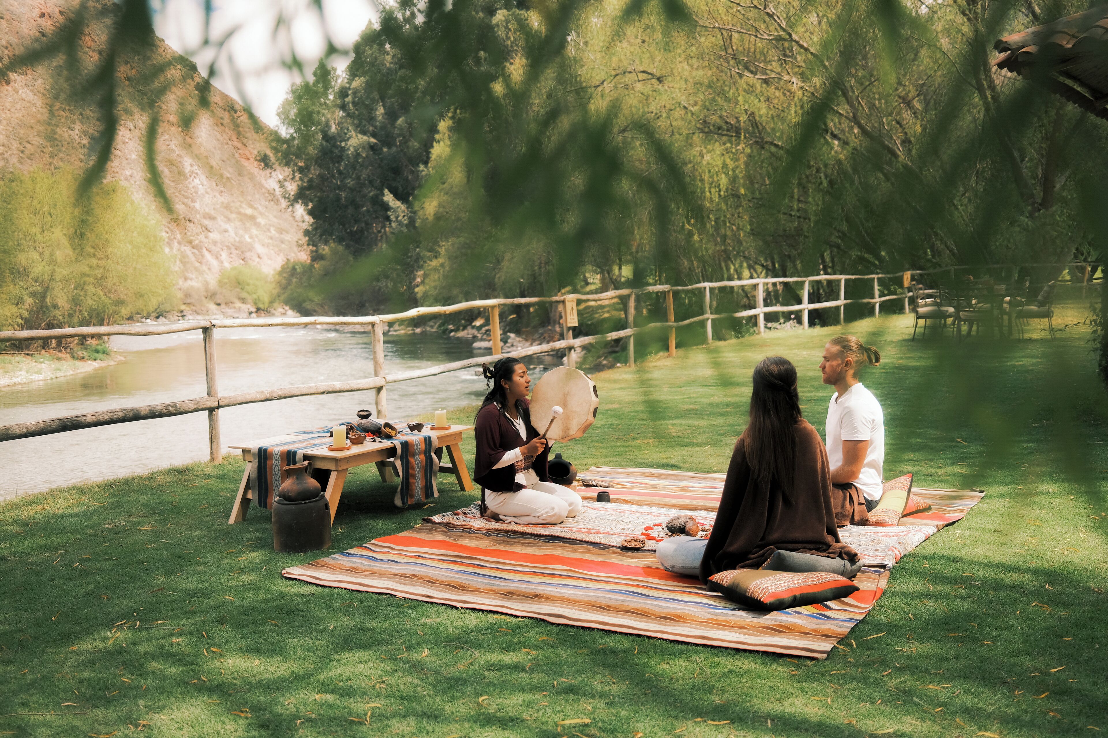 A group of people sitting on a striped blanket outdoors near a river, with a wooden fence and trees in the background.