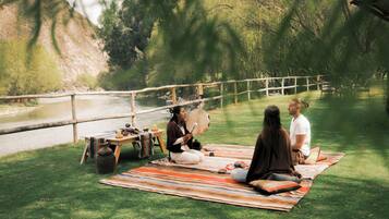 A group of people sitting on a striped blanket outdoors near a river, with a wooden fence and trees in the background.