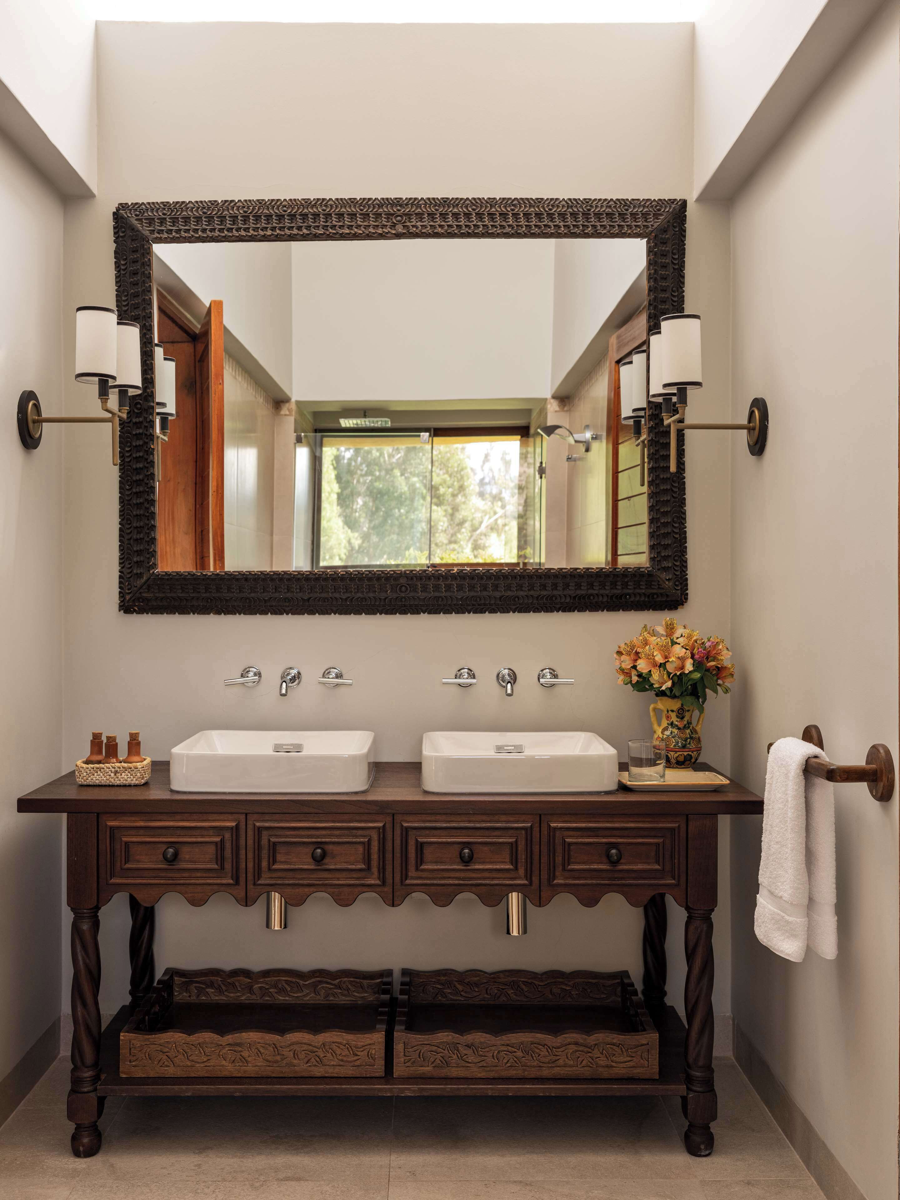 A bathroom with two sinks, a large mirror, wall sconces, a vase with flowers, and a towel rack.